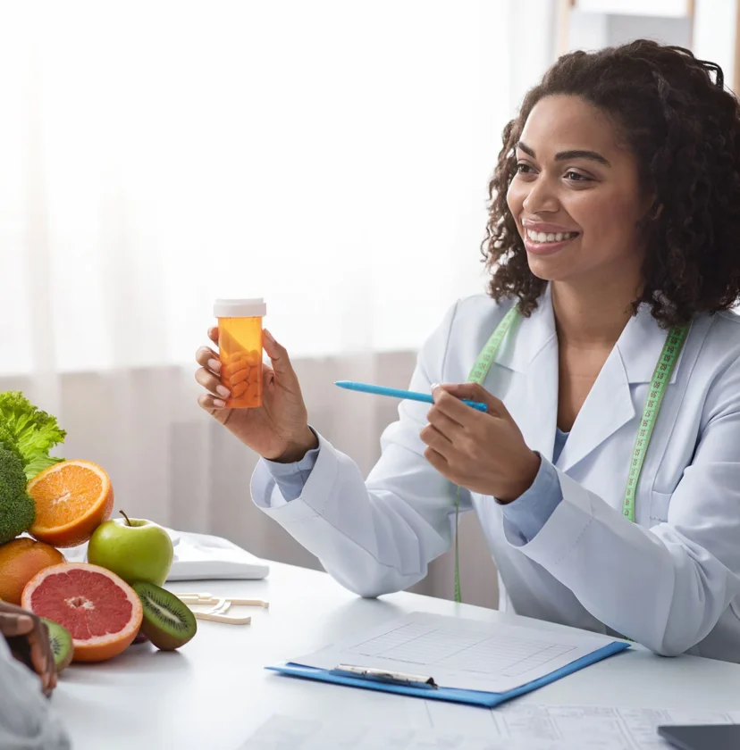 A doctor holding a pill bottle and pen sits across from a patient. Fresh fruits and vegetables are on the table.