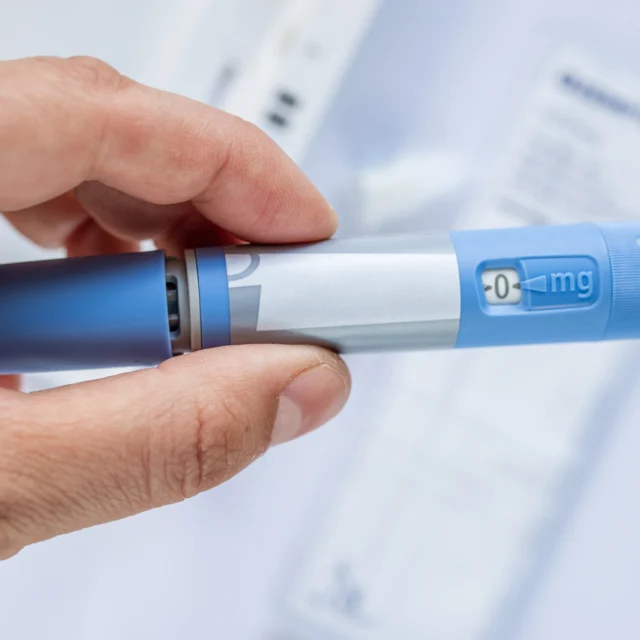 A hand holds a blue and silver insulin pen with a dosage dial set to 0 mg.