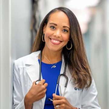 A woman in a white coat and blue shirt stands smiling, holding a stethoscope around her neck. She has long hair and wears a necklace and smartwatch.