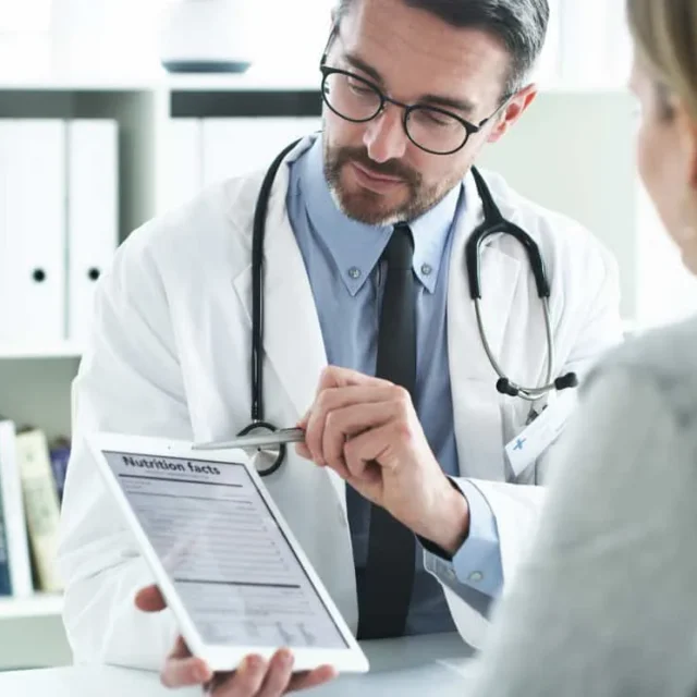 Doctor in a white coat shows a tablet screen to a seated woman in an office setting, with books and binders in the background.