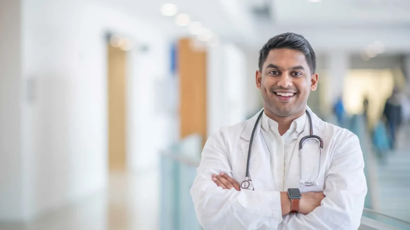 A doctor in a white coat with a stethoscope around his neck stands smiling in a bright hospital hallway.